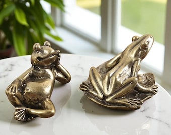 Könnte beinhalten: Zwei Froschfiguren aus Bronze. Ein Frosch posiert mit dem Kinn auf der Hand, der andere liegt auf einem Blatt. Die Frösche befinden sich auf einer weißen Marmoroberfläche, mit einer grünen Pflanze und einem Fenster im Hintergrund.
