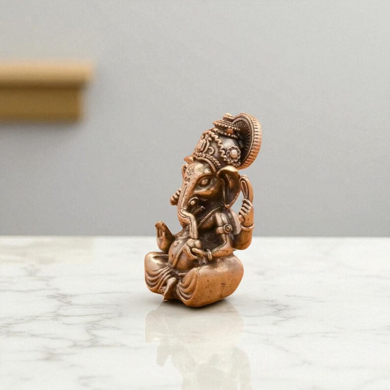 May include: A bronze-colored statue of the Hindu deity Ganesha, seated in a meditative pose. The statue features intricate details, including a large head with elephant features, ornate headdress, and multiple arms. The sculpture is on a marble surface.
