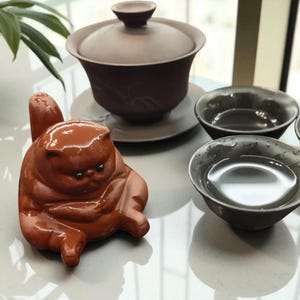 May include: A brown ceramic tea set with a teapot, lid, saucer, and two small teacups. A brown ceramic cat figurine is in the foreground. The tea set is a muted brown colour.