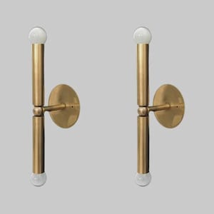 May include: Two brass wall sconces with a minimalist design. Each sconce features a vertical rod with a bulb at each end, connected to a circular wall mount. The sconces have a warm, metallic finish and a modern aesthetic.