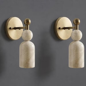 May include: A pair of wall sconces featuring a circular brushed brass backplate, a horizontal arm with a spherical finial, and a vertical pendant component consisting of a stacked marble sphere and a cylindrical marble shade in a beige tone.