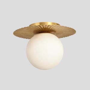 May include: A modern ceiling light fixture featuring a spherical, off-white glass globe. The globe is suspended beneath a decorative, scalloped brass plate. The fixture has a minimalist design, suitable for various interior styles.
