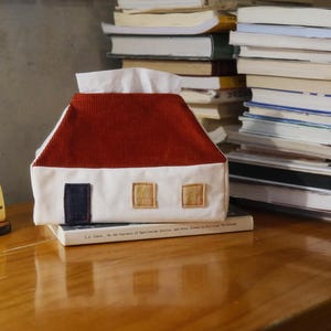 Puede incluir: Una funda para caja de pañuelos con forma de casa con tejado de pana rojo y paredes blancas. La casa tiene una puerta azul oscuro y dos ventanas cuadradas color beige. Libros y un juguete VW bus en el fondo.