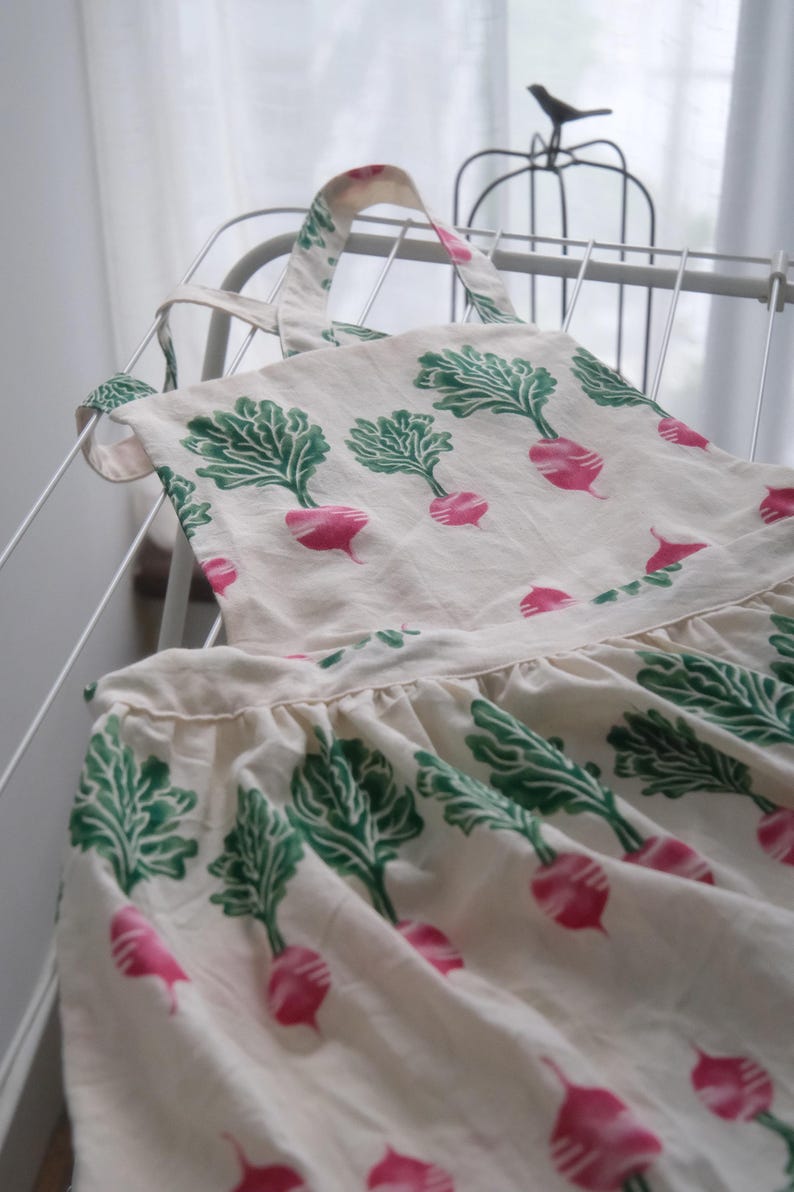 Puede incluir: Delantal blanco roto con un estampado de r&aacute;banos rosas y hojas verdes. El delantal tiene una falda con volantes y correas ajustables. Cuelga de un estante blanco.