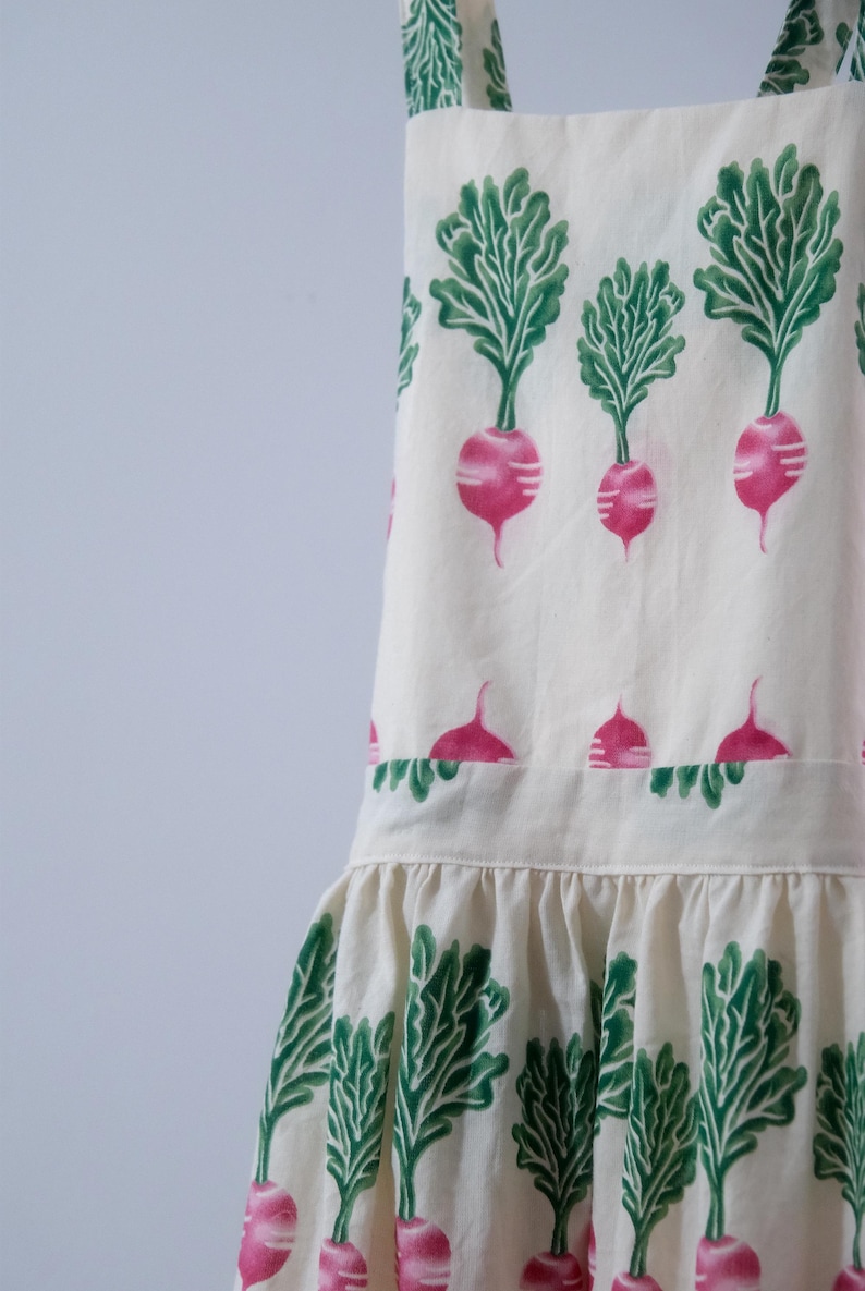 Puede incluir: Delantal blanco roto con un estampado repetido de r&aacute;banos rosas y hojas verdes. El delantal tiene un dobladillo con volantes y correas, ideal para la cocina o el jard&iacute;n. El dise&ntilde;o de r&aacute;bano a&ntilde;ade un toque de fantas&iacute;a.