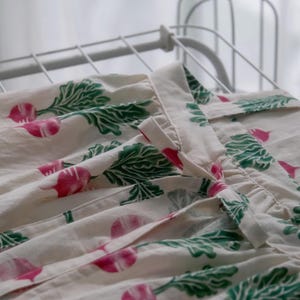Puede incluir: Tela color crema con un estampado repetido de r&aacute;banos rosas y hojas verdes. La tela est&aacute; drapeada sobre un tendedero blanco. Los r&aacute;banos tienen una raya blanca en el centro.