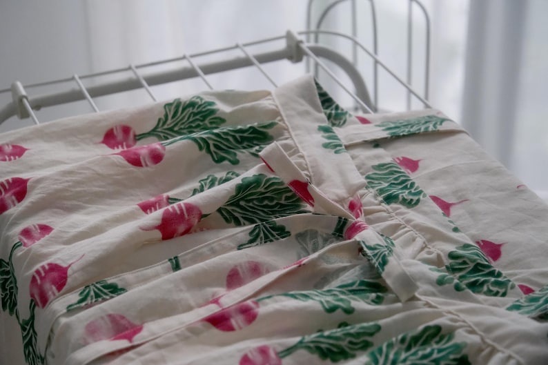 Puede incluir: Tela color crema con un estampado repetido de r&aacute;banos rosas y hojas verdes. La tela est&aacute; drapeada sobre un tendedero blanco. Los r&aacute;banos tienen una raya blanca en el centro.