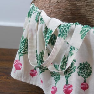 Puede incluir: Un bolso tote blanco con un patr&oacute;n repetido de r&aacute;banos rosas y hojas verdes. El bolso tiene un dise&ntilde;o sencillo con un asa y est&aacute; parcialmente dentro de una cesta de mimbre. El bolso est&aacute; hecho de tela clara y se encuentra sobre una superficie de madera.