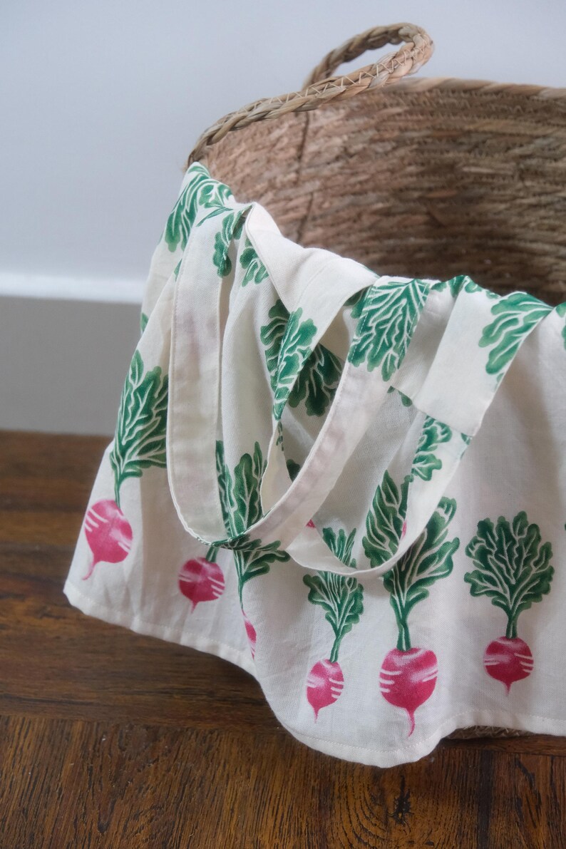 Puede incluir: Un bolso tote blanco con un patr&oacute;n repetido de r&aacute;banos rosas y hojas verdes. El bolso tiene un dise&ntilde;o sencillo con un asa y est&aacute; parcialmente dentro de una cesta de mimbre. El bolso est&aacute; hecho de tela clara y se encuentra sobre una superficie de madera.