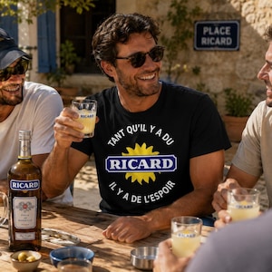Peut inclure: Un t-shirt noir à manches courtes avec un motif de soleil jaune, un logo rectangulaire bleu avec le texte blanc RICARD, et le texte circulaire blanc TANT QU'IL Y A DU RICARD IL Y A DE L'ESPOIR imprimé sur la poitrine.