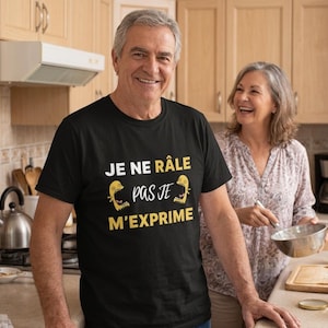 Peut inclure: Un t-shirt noir avec le texte français "JE NE RÂLE PAS JE M'EXPRIME" en blanc et jaune. Le t-shirt présente un dessin de créature jaune. Un homme porte le t-shirt dans une cuisine.