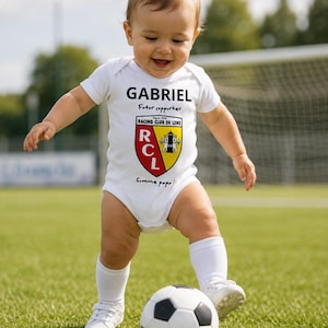 Peut inclure: Un body blanc à manches courtes pour bébé avec le prénom GABRIEL en noir, la mention Futur supporter, le logo du Racing Club de Lens aux couleurs rouge, jaune et noir, et l'inscription Comme papa imprimés sur le devant de la poitrine.