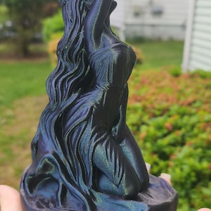 May include: A three-dimensional sculpture of a kneeling figure with long, flowing hair, rendered in a color-shifting filament that transitions between deep purple, dark blue, and metallic green hues, set upon a circular, textured base.