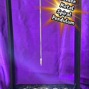 May include: A black wooden pendulum stand with a golden metal spiral pendulum suspended from the top. The base of the stand has a celestial design. The image includes the text "Golden Metal Spiral Pendulum".