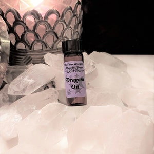 May include: A small brown glass bottle with a black cap, labeled "Dragons Oil" and "The Three Wise Cats Magical Shoppe". The bottle is sitting on a bed of white crystal rocks.