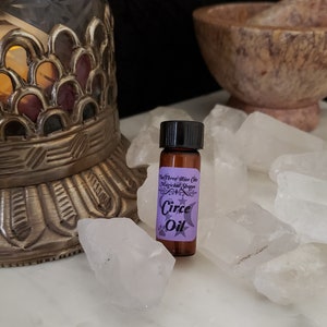 May include: A small brown glass bottle with a black cap, labeled "The Three Wise Cats Magical Shoppe Circe Oil" on a purple label with a crescent moon and stars. The bottle is surrounded by white crystal rocks.