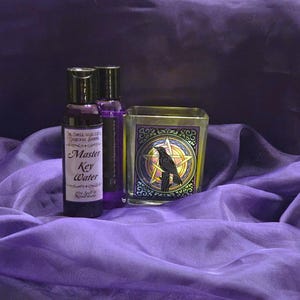 Puede incluir: Una colección de artículos rituales sobre una tela morada. Los artículos incluyen dos botellas de Master Key Water y un portavelas de vidrio con un diseño de cuervo y pentagrama. Las botellas son de color púrpura oscuro y el portavelas tiene un borde dorado.