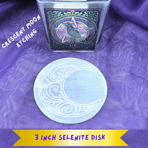 May include: A 3 inch selenite disk with a crescent moon etching. The disk is white and the etching is in a light gray color. The disk is on a purple fabric background.