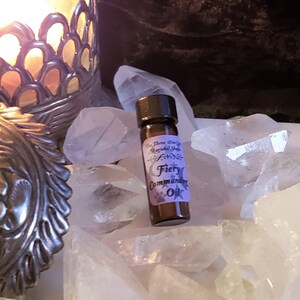 May include: A small brown glass bottle with a black cap, containing a clear liquid. The bottle has a white label with the text "The Three Wise Co. Magical Shoppe Fiery Commanding Oil". The bottle is sitting on a bed of white crystals.