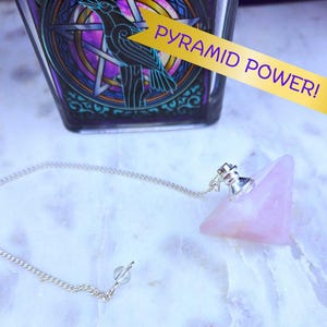 May include: A pink crystal pyramid pendulum with a silver chain and a silver top. The pendulum is hanging from a chain and is resting on a white surface. The text "PYRAMID POWER!" is visible in the background.
