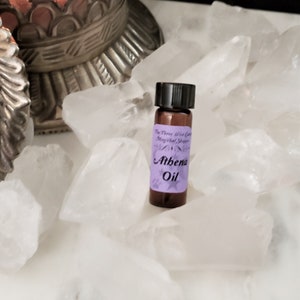 May include: A small brown glass bottle with a black cap and a purple label that reads "The Three Wise Cats Magical Shoppe" and "Athena Oil". The bottle is sitting on a bed of white quartz crystals.