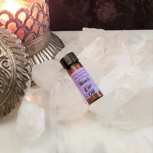 May include: A small brown glass bottle with a purple label that reads "The Three Wise Cat Magical Shoppe Black Cat Oil". The bottle is sitting on a bed of white crystal rocks.