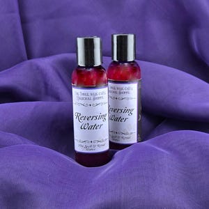 May include: Two small bottles of purple liquid labelled "Reversing Water" with the text "The Three Wise Cats Magickal Shoppe" on the front. The bottles are sitting on a purple fabric surface.