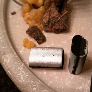 May include: Two charcoal sticks, one silver and one black, on a light brown surface. The sticks are rectangular and have a slightly rounded edge.