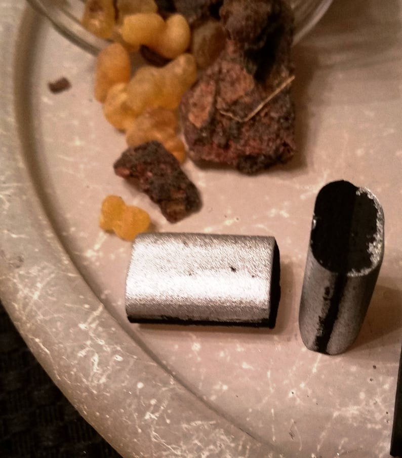 May include: Two charcoal sticks, one silver and one black, on a light brown surface. The sticks are rectangular and have a slightly rounded edge.