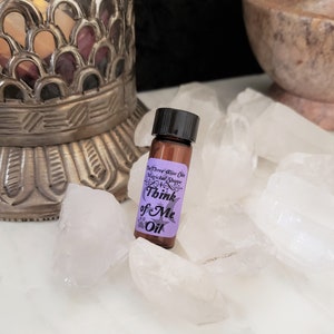 Può includere: Un piccolo flacone di vetro marrone con un tappo nero e un'etichetta viola che recita "The Three Wise Cats Mystical Shoppe Think of Me Oil". Il flacone è appoggiato su un letto di cristalli bianchi.