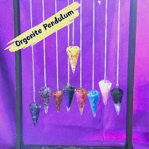 May include: A display of seven orgonite pendulums in various colours, suspended from a black wooden stand. The pendulums are cone-shaped and hang from silver chains. The text "Orgonite Pendulum" is displayed on the image.