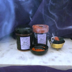 Könnte beinhalten: Anordnung von Räucherwerkprodukten. Zwei Gläser und eine kleine Schale mit rotbraunem Pulver sind zu sehen, beschriftet mit "Road Opener Powder Incense". Ein kleiner, goldfarbener Räuchergefäß mit brennendem Räucherwerk gibt Rauch ab. Der Hintergrund ist dunkelblau.