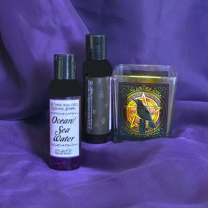 May include: Three bottles and a candle on a purple surface. One bottle is labelled "Ocean/Sea Water". The candle has a black raven design inside a star. The bottles are dark with black caps.