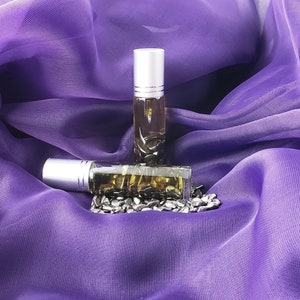 May include: Two small, clear glass roll-on bottles with silver caps, filled with a dark liquid. One bottle is upright, resting on top of the other. The bottles are on a bed of small, dark stones, against a backdrop of draped purple fabric.