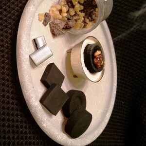 May include: A white ceramic plate with a silver incense burner, black charcoal squares, and a small pile of brown and yellow resin. The incense burner has a black charcoal disc with a brown resin piece on top. Smoke is rising from the burner.