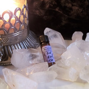 May include: A small brown glass bottle with a purple label that reads "The Three Wise Men Magical Shop Evil Eye Oil". The bottle is sitting on a bed of white crystal points.