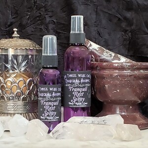 May include: Two purple glass bottles with spray tops labeled "Three Wise Co. Hagickal Shoppe Tranquil Rest Spray" on a white surface with clear quartz crystals and a brown marble mortar and pestle.