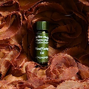 Könnte beinhalten: Eine kleine, dunkelgrüne Glasflasche mit goldenem Deckel. Die Flasche ist mit "The Three Wise On Magical Shoppe Hand Fasting Oil" beschriftet.