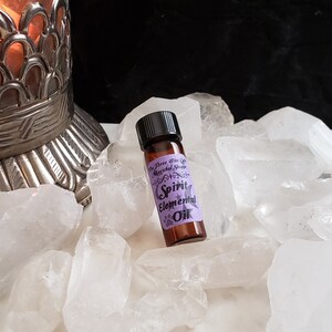May include: A small brown glass bottle with a black cap, labeled "Spirit Elemental Oil" with a purple background and a white star. The bottle is sitting on a bed of white quartz crystals.