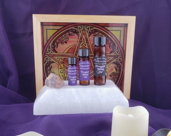 May include: Three small bottles of Dragon's Blood Oil, with a purple label and a gold pentagram design. The bottles are on a white surface in front of a wooden frame with a stained glass pentagram design.