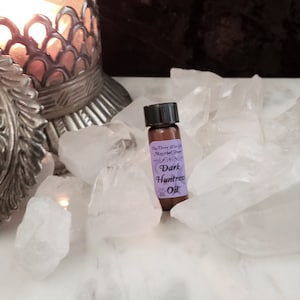 May include: A small brown glass bottle with a black cap and a purple label that reads "The Three Wise Co. Mystical Supplies Dark Huntress Oil". The bottle is sitting on a bed of white quartz crystals.