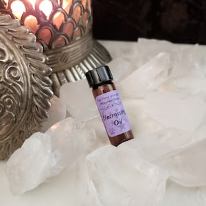 May include: A small brown glass bottle with a black cap, labelled "The Three Wise Co. Magical Shoppe Uncrossing Oil". The bottle is sitting on a bed of white crystals.