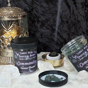 May include: A display of "Commanding Incense Powder" jars from "The Three Wise Cats Magickal Shoppe." One jar is open, showing green incense. A decorative metal container and crystals are also present, with smoke rising from a small incense holder.