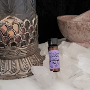 May include: A small brown glass bottle with a black cap and a purple label that reads "The Three Wise One Magical Shoppe Spellbound Oil". The bottle is sitting on a bed of white crystals.