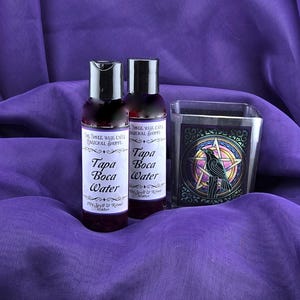 Könnte beinhalten: Zwei durchsichtige Glasflaschen mit schwarzen Kappen und Etiketten mit der Aufschrift "Three Wise Cats Magical Shoppe" und "Tapa Boca Water 20% Spell & Ritual Water". Die Flaschen stehen auf einem violetten Stoff neben einem Glaskerzenhalter mit einem Buntglasdesign eines schwarzen Rabens in einem fünfzackigen Stern.