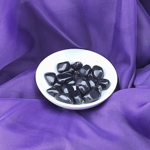Könnte beinhalten: Eine weiße Schale gefüllt mit schwarzen, abgerundeten Steinen. Die Schale steht auf einem violetten Stoffhintergrund.