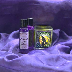 May include: Two purple bottles and a glass candle holder with a raven and pentagram design. The bottles have black caps and labels that read "Dominate All Water". The candle holder has a gold rim and a black raven illustration.