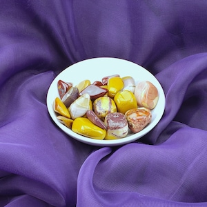 May include: A white bowl filled with a variety of polished stones in shades of yellow, brown, and red. The stones are arranged in a random pattern and are surrounded by a purple fabric background.