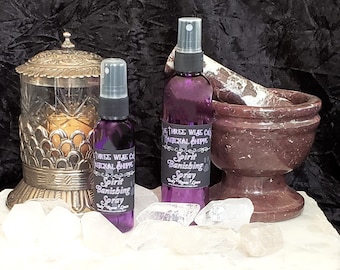 May include: Two purple glass bottles with spray tops and black labels that read "Spirit Banishing Spray" on a white surface with clear quartz crystals. The bottles are next to a brown mortar and pestle and a gold decorative stand with a glass top.