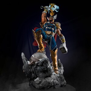 Op de afbeelding: Een gedetailleerd beeld van een superheld in een dynamische pose, gekleed in blauwe en gouden bepantsering met een rode cape. De figuur houdt een gloeiende hamer en een wapen vast, staand op een rotsachtige basis. Het beeld is ongeveer 30 cm hoog.